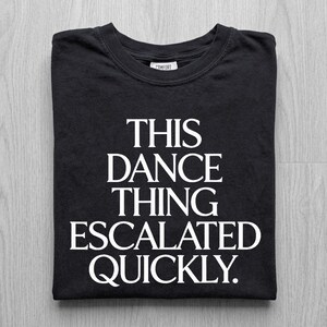 May include: Black t-shirt with the white text "THIS DANCE THING ESCALATED QUICKLY." The shirt is folded neatly, showcasing the design. The shirt has a crew neck and a tag that says "COMFORT".
