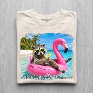May include: Cream-colored t-shirt featuring a raccoon wearing heart-shaped sunglasses and floating on a pink flamingo float in the ocean. The raccoon holds a cocktail. The words "feral girl summer" are printed below the image.