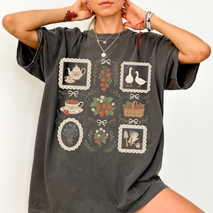 May include: Dark gray t-shirt featuring a charming garden-themed design. The design includes framed illustrations of a teapot, teacup, strawberries, geese, a basket, gardening tools, and flowers. The shirt has a relaxed fit.