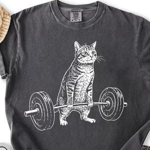 May include: Dark gray t-shirt featuring a white illustration of a cat lifting a barbell. The cat is depicted in a strong pose, with the barbell held in front of it. The shirt has a crew neck and short sleeves. Black sneakers and blue jeans are visible.