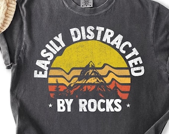 Puede incluir: Camiseta gris oscuro con el texto blanco "EASILY DISTRACTED BY ROCKS". El diseño presenta una puesta de sol amarilla y naranja con una silueta de montaña. Prenda de vestir informal.