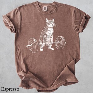 May include: Espresso-colored t-shirt featuring a white graphic of a cat lifting a barbell. The cat is drawn in detail, with stripes and whiskers. The shirt has a crew neck and short sleeves. The text "Espresso" is at the bottom of the shirt.