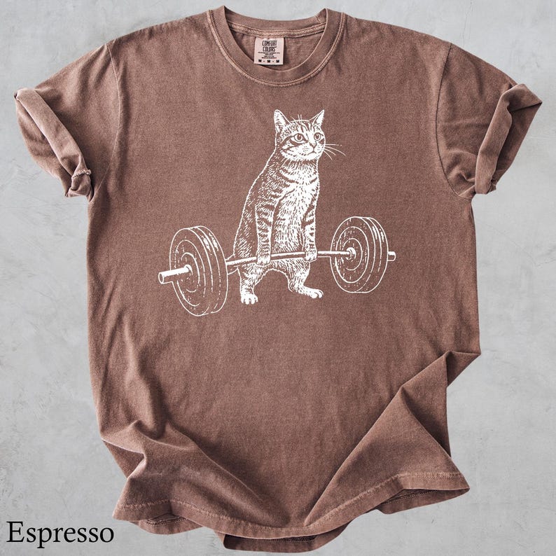 May include: Espresso-colored t-shirt featuring a white graphic of a cat lifting a barbell. The cat is drawn in detail, with stripes and whiskers. The shirt has a crew neck and short sleeves. The text "Espresso" is at the bottom of the shirt.