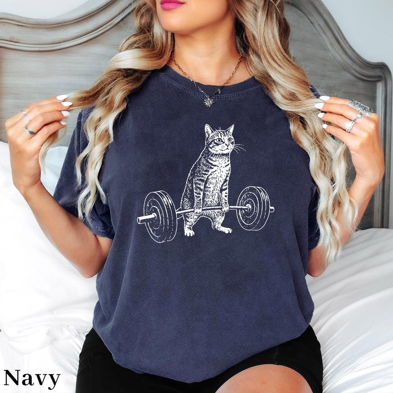 May include: Navy blue t-shirt featuring a white graphic of a cat lifting a barbell. The cat is depicted in a detailed, realistic style. The word "Navy" is printed in white at the bottom left of the shirt.