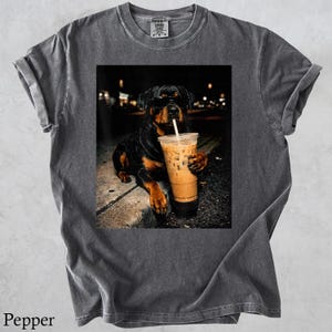 May include: Dark gray t-shirt with a graphic of a Rottweiler wearing sunglasses and drinking iced coffee. The dog is holding the coffee cup with its paw. The shirt has a crew neck and short sleeves.