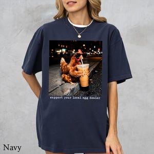 May include: A navy blue t-shirt featuring a graphic of a brown chicken wearing black sunglasses and holding a plastic cup with a straw. Below the image, the text reads, support your local egg dealer. The shirt has short sleeves and a crew neckline.