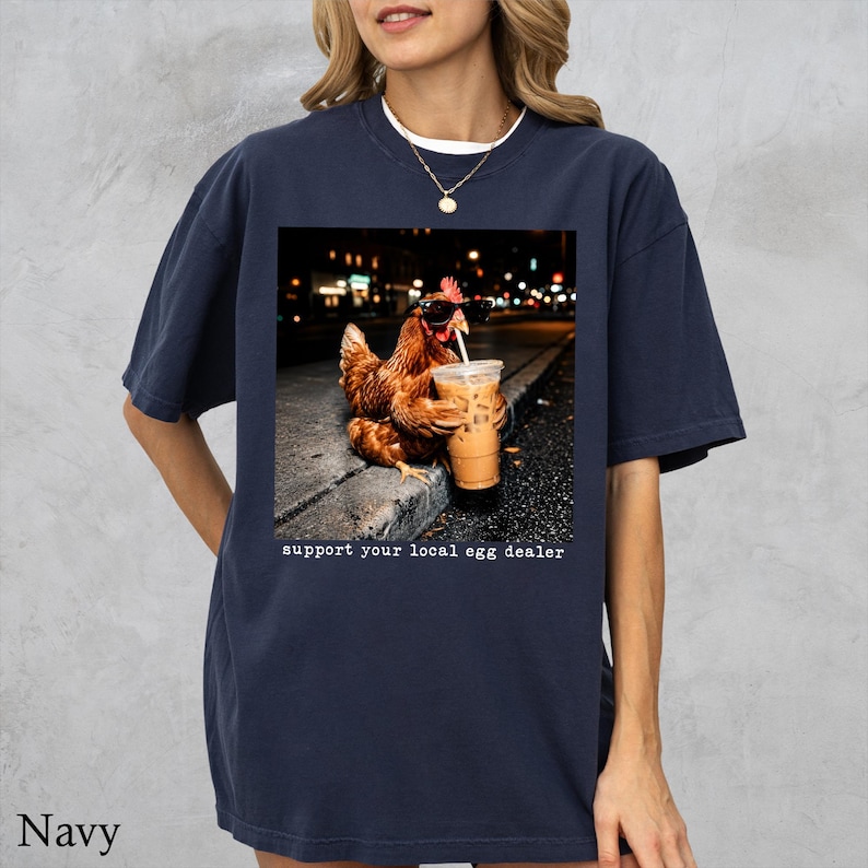 May include: A navy blue t-shirt featuring a graphic of a brown chicken wearing black sunglasses and holding a plastic cup with a straw. Below the image, the text reads, support your local egg dealer. The shirt has short sleeves and a crew neckline.