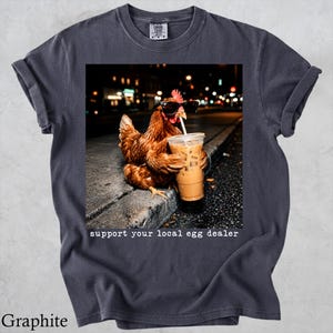 May include: A graphite gray t-shirt featuring a graphic of a brown chicken wearing black sunglasses and drinking an iced coffee from a plastic cup with a straw. The text below the image reads, support your local egg dealer.