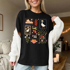 Pode incluir: T-shirt preto com um design caprichoso com ilustra&ccedil;&otilde;es emolduradas de um ganso, morangos, flores e uma chaleira. O design inclui um ganso usando um chap&eacute;u, uma cesta de morangos e um buqu&ecirc; de flores. A camisa &eacute; um item de roupa casual.