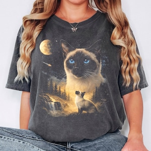 May include: A charcoal grey short-sleeve t-shirt featuring a graphic print of a Siamese cat with bright blue eyes, a smaller Siamese cat sitting in a forest landscape with a waterfall, a glowing moon, and a shooting star in a starry night sky.