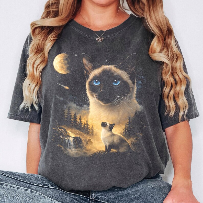 May include: A charcoal grey short-sleeve t-shirt featuring a graphic print of a Siamese cat with bright blue eyes, a smaller Siamese cat sitting in a forest landscape with a waterfall, a glowing moon, and a shooting star in a starry night sky.