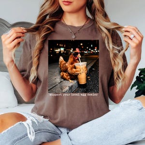 May include: A brown short-sleeve t-shirt featuring a graphic of a chicken wearing black sunglasses and holding an iced coffee with a straw. Below the image, the text reads, support your local egg dealer. The shirt has a crew neckline and a relaxed fit.