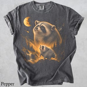 May include: A charcoal gray t-shirt featuring a graphic print of a large raccoon looking up at a crescent moon and a smaller raccoon standing on a rock near a waterfall. The design includes golden yellow highlights, stars, and a shooting star.