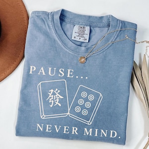 May include: A blue short-sleeved t-shirt with a crew neckline and a Comfort Colors label. The front features the white text PAUSE... NEVER MIND. and a graphic of two mahjong tiles, one with a Chinese character and one with six circular dots.