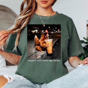 May include: A dark olive green t-shirt featuring a graphic of a brown chicken wearing black sunglasses and holding a plastic cup with a straw. The text "support your local egg dealer" is printed in white letters below the chicken graphic.