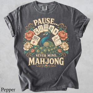 May include: A charcoal gray short-sleeve t-shirt featuring a graphic print of a peacock surrounded by colorful flowers and mahjong tiles. The text reads PAUSE... NEVER MIND, MAHJONG. The design includes intricate gold and floral accents.