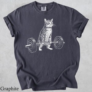 May include: Graphite-colored t-shirt featuring a white illustration of a cat lifting a barbell. The cat is depicted with detailed stripes and a focused expression. The barbell has circular weights on each end.