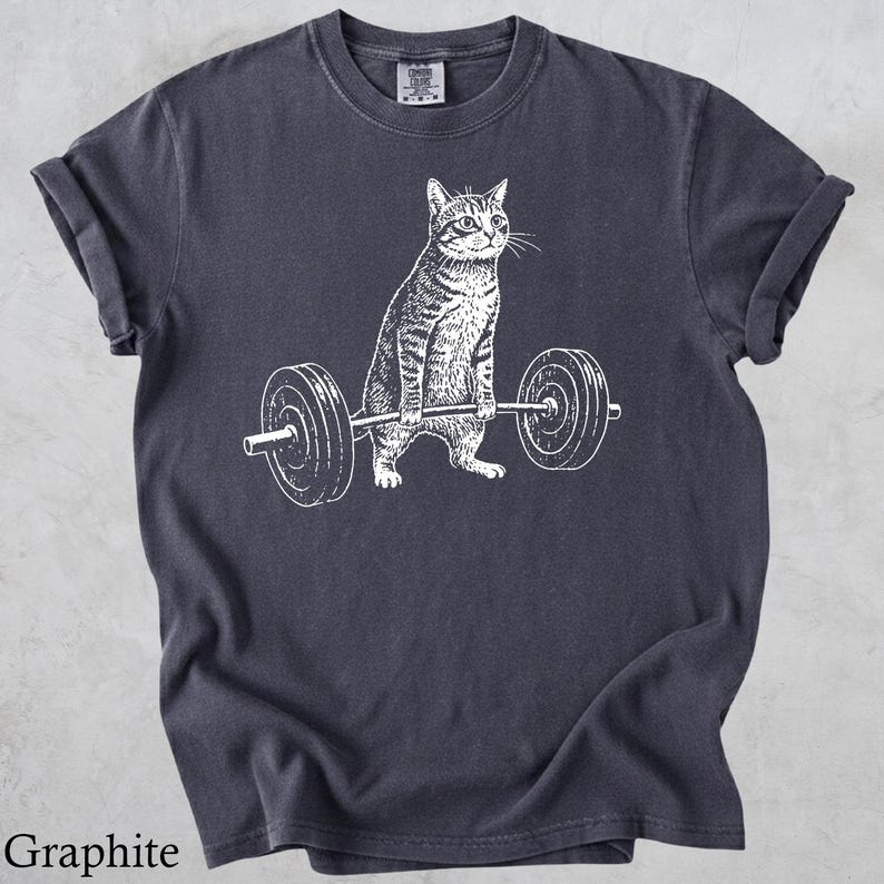 May include: Graphite-colored t-shirt featuring a white illustration of a cat lifting a barbell. The cat is depicted with detailed stripes and a focused expression. The barbell has circular weights on each end.