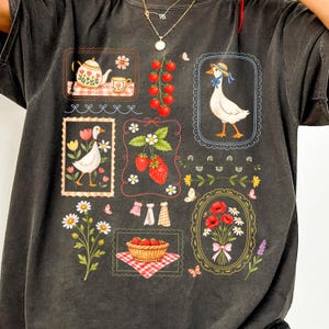 Pode incluir: Camiseta cinza escuro com uma colagem de ilustra&ccedil;&otilde;es. O design inclui uma chaleira, morangos, um ganso usando um chap&eacute;u, flores e uma cesta de morangos. As ilustra&ccedil;&otilde;es s&atilde;o emolduradas com bordas decorativas.