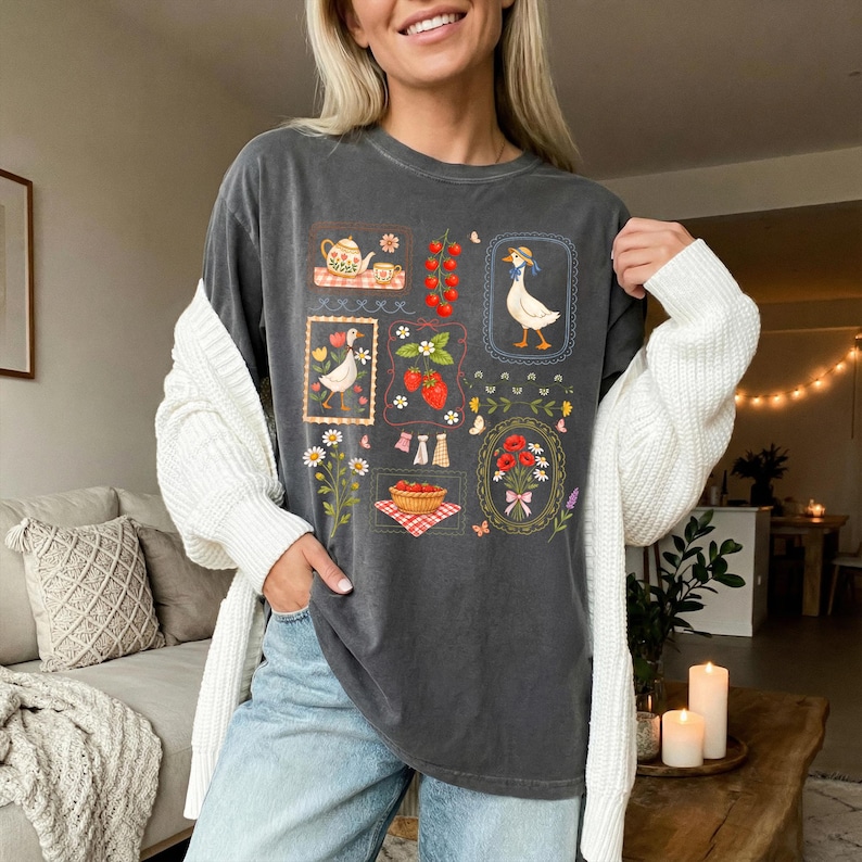 Pode incluir: T-shirt cinzenta escura com um design caprichoso com ilustra&ccedil;&otilde;es emolduradas de um ganso, uma chaleira, morangos e flores. A camisa tem um ajuste descontra&iacute;do e &eacute; feita de material macio. O design inclui vermelho, branco e verde.