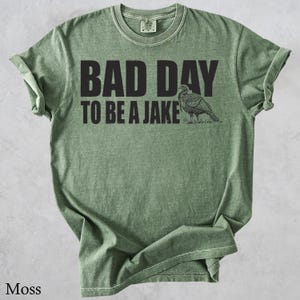May include: A moss green t-shirt with the text "BAD DAY TO BE A JAKE" in black block letters. A detailed illustration of a turkey is to the right of the text. The shirt has a crew neck and short sleeves.