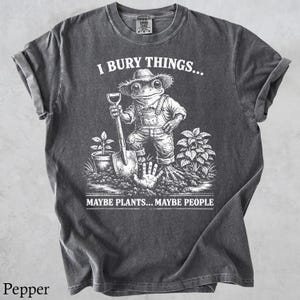 May include: Pepper-colored t-shirt with a humorous graphic. The design features a frog in overalls and a straw hat holding a shovel, with the text "I BURY THINGS... MAYBE PLANTS... MAYBE PEOPLE."