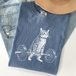 May include: A blue t-shirt with a white graphic of a cat lifting a barbell. The cat is drawn in a detailed, realistic style. The shirt is folded on top of a pair of blue jeans and a tan hat.