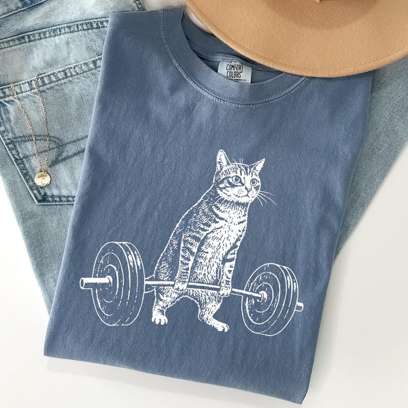 May include: A blue t-shirt with a white graphic of a cat lifting a barbell. The cat is drawn in a detailed, realistic style. The shirt is folded on top of a pair of blue jeans and a tan hat.