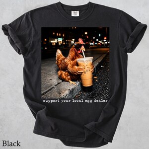 May include: A black t-shirt featuring a graphic of a brown chicken wearing black sunglasses and holding a plastic cup of iced coffee with a straw. The text below the image reads, "support your local egg dealer" in a white typewriter-style font.