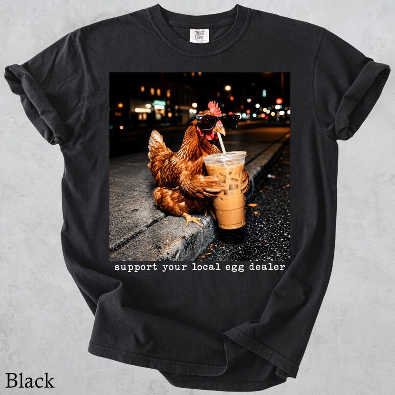 May include: A black t-shirt featuring a graphic of a brown chicken wearing black sunglasses and holding a plastic cup of iced coffee with a straw. The text below the image reads, "support your local egg dealer" in a white typewriter-style font.