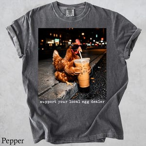 May include: A charcoal grey t-shirt featuring a graphic of a brown chicken wearing black sunglasses and holding a plastic cup with a straw. Below the image, the text reads, support your local egg dealer. The shirt has a crew neck and short sleeves.