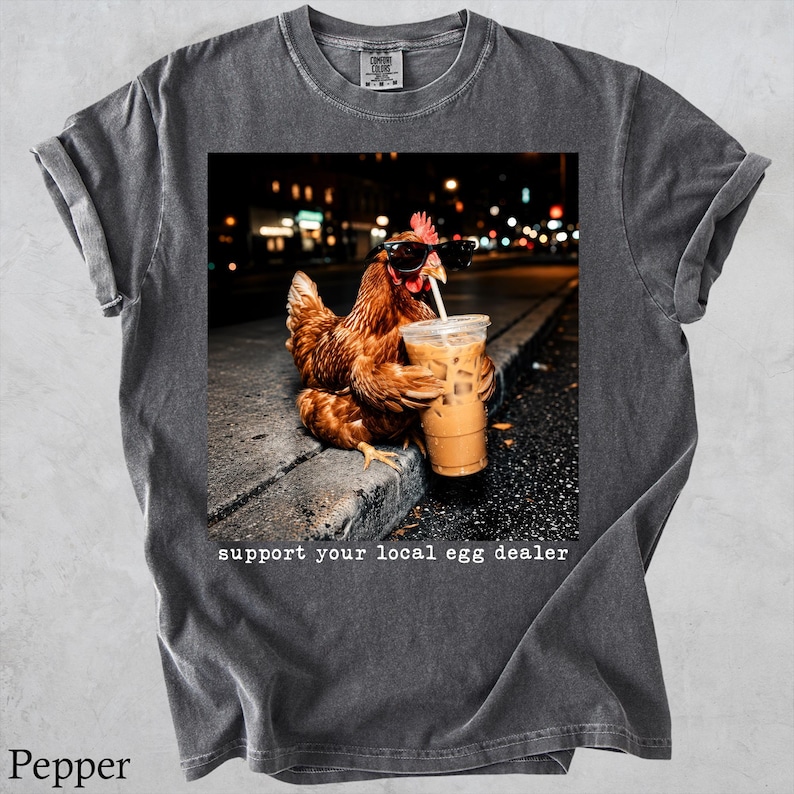 May include: A charcoal gray t-shirt featuring a graphic of a brown chicken wearing black sunglasses and holding a plastic cup with a straw. Below the image, the text reads, support your local egg dealer. The shirt has a crew neck and short sleeves.