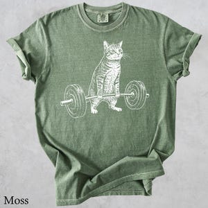 May include: Moss green t-shirt featuring a white graphic of a cat lifting a barbell. The cat is drawn in a detailed, realistic style. The shirt is made of a soft material and has a classic crew neck.