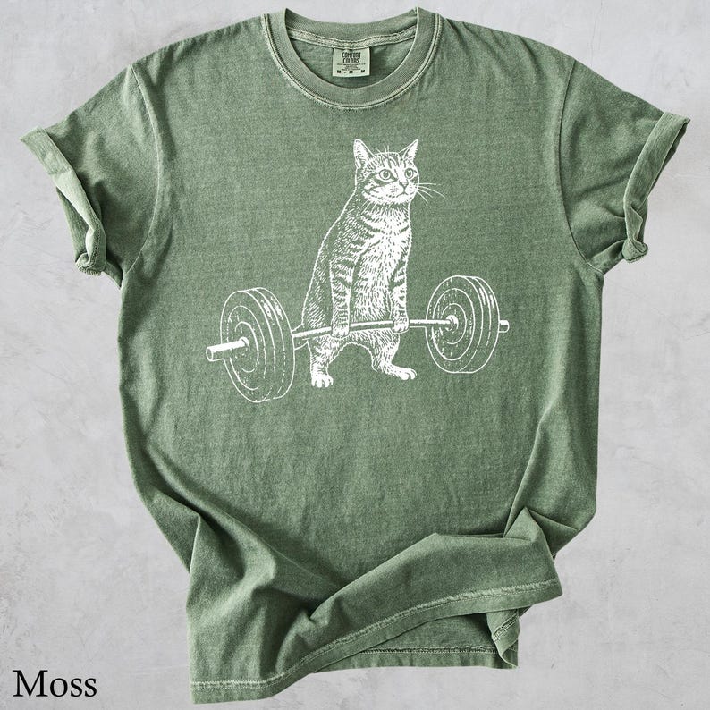 May include: Moss green t-shirt featuring a white graphic of a cat lifting a barbell. The cat is drawn in a detailed, realistic style. The shirt is made of a soft material and has a classic crew neck.