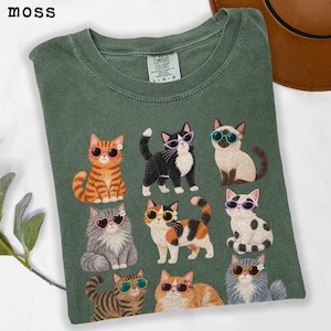 May include: A moss green crew neck t-shirt featuring a graphic print of nine cats wearing colorful flower-shaped sunglasses. The cats are depicted in various breeds and colors, including orange tabby, black and white, and calico, arranged in a grid pattern on the front of the garment.