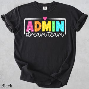 May include: A black short-sleeved t-shirt featuring the text ADMIN in colorful, polka-dotted block letters, with the word dream team written in white cursive below. A small pink heart sits above the text, all enclosed in a white rectangular frame.