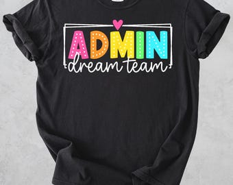 May include: A black short-sleeved t-shirt featuring the text ADMIN in colourful, polka-dotted block letters, with the word dream team written in white cursive below. A small pink heart sits above the text, all enclosed in a white rectangular frame.
