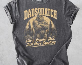 May include: Dark grey t-shirt featuring a Bigfoot-style illustration and the words "Dadsquatch". The design includes the phrase "Like a Regular Dad, Just More Squatchy" in a circular design.