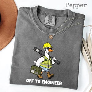 May include: Gray t-shirt with a cartoon duck wearing a yellow hard hat, sunglasses, and a safety vest, carrying a blueprint and toolbox. The text "OFF TO ENGINEER" is printed at the bottom.