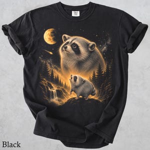 May include: A black t-shirt featuring a graphic print of a large raccoon looking up at a glowing moon and a smaller raccoon standing on a rock near a waterfall in a forest setting. The design includes a starry night sky with a shooting star.