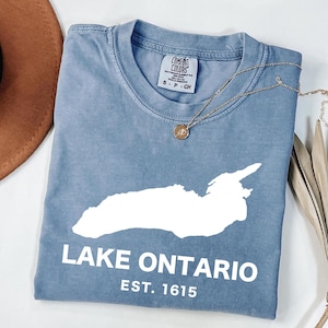 May include: A blue short-sleeved t-shirt featuring a white graphic of Lake Ontario, the text "LAKE ONTARIO" and "EST. 1615" in white lettering. The shirt has a crew neckline and a Comfort Colors brand label printed on the inside of the collar.
