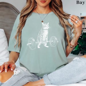 May include: A light green t-shirt featuring a white graphic of a cat lifting a barbell. The cat is depicted in a strong pose, with the barbell held high. The shirt is a relaxed fit, suitable for casual wear.