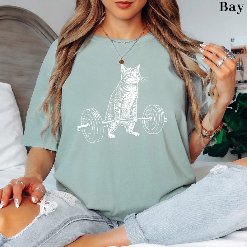 May include: A light green t-shirt featuring a white graphic of a cat lifting a barbell. The cat is depicted in a strong pose, with the barbell held high. The shirt is a relaxed fit, suitable for casual wear.