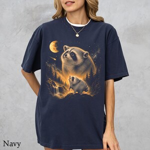 May include: A navy blue t-shirt featuring a graphic print of two raccoons in a forest setting at night. The design includes a large moon, a shooting star, and a waterfall, all rendered in warm golden and brown tones against the dark fabric.