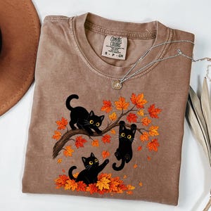 May include: A brown short-sleeve t-shirt featuring a graphic of three black cats playing in a tree with vibrant orange and red autumn leaves. The shirt has a crew neckline and a Comfort Colors brand label visible on the inside of the collar.