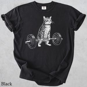 May include: Black t-shirt featuring a white graphic of a cat lifting a barbell. The cat is depicted in a strong, athletic pose, with the barbell resting on its paws. The text "Black" is at the bottom of the shirt.