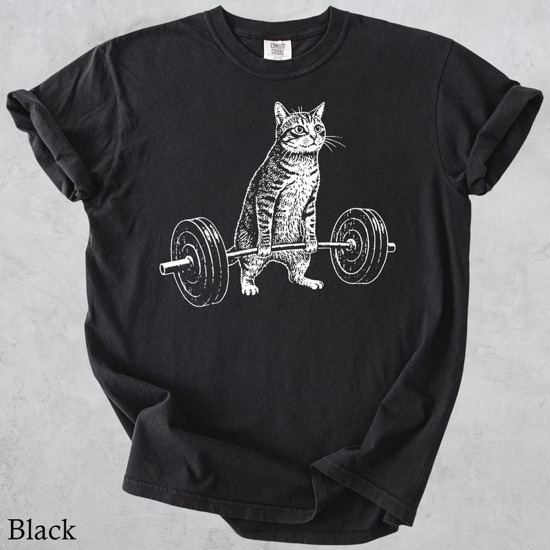 May include: Black t-shirt featuring a white graphic of a cat lifting a barbell. The cat is depicted in a strong, athletic pose, with the barbell resting on its paws. The text "Black" is at the bottom of the shirt.