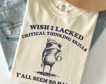 May include: An ivory t-shirt featuring a dark blue graphic of a frog wearing a beanie and holding a mug. The text reads: WISH I LACKED CRITICAL THINKING SKILLS Y'ALL SEEM SO HAPPY. The shirt has a Comfort Colors label at the inner neckline.