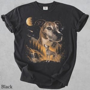 May include: A black short-sleeved t-shirt featuring a graphic print of a dog's face in the night sky above a smaller dog standing on a rock near a waterfall and pine trees. The scene includes a glowing moon and stars in shades of gold and brown.