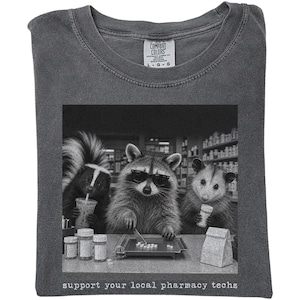 May include: A charcoal gray t-shirt featuring a black and white graphic of a skunk, a raccoon wearing sunglasses, and an opossum behind a pharmacy counter. The animals are handling pill bottles and tablets. The text reads: support your local pharmacy techs.